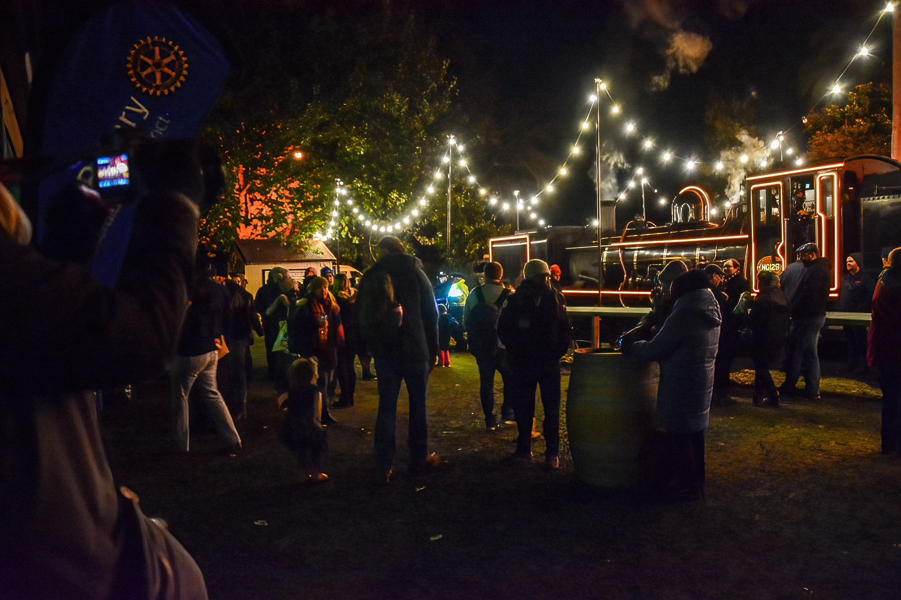 An outdoor area lit with fairy lights. A number of people are standing around the space. A steam locomotive can be seen in the background in front of a lit up water tank.