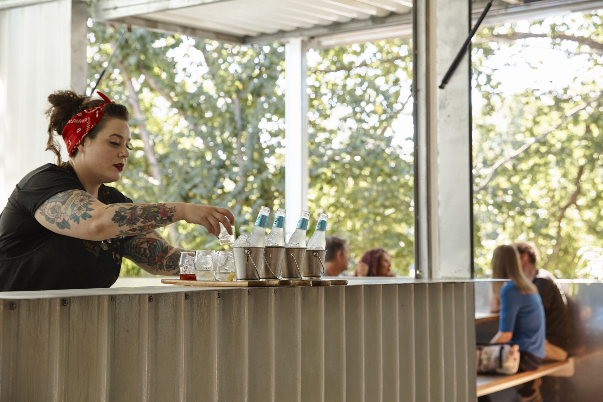 A photo of a bartender at a counter filling glasses with gin. Patrons can be seen sitting at a table in the background.