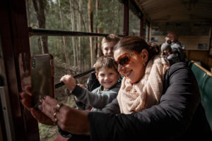 A woman and two children inside an open side train carriage. They are taking a selfie and are smiling at the camera.