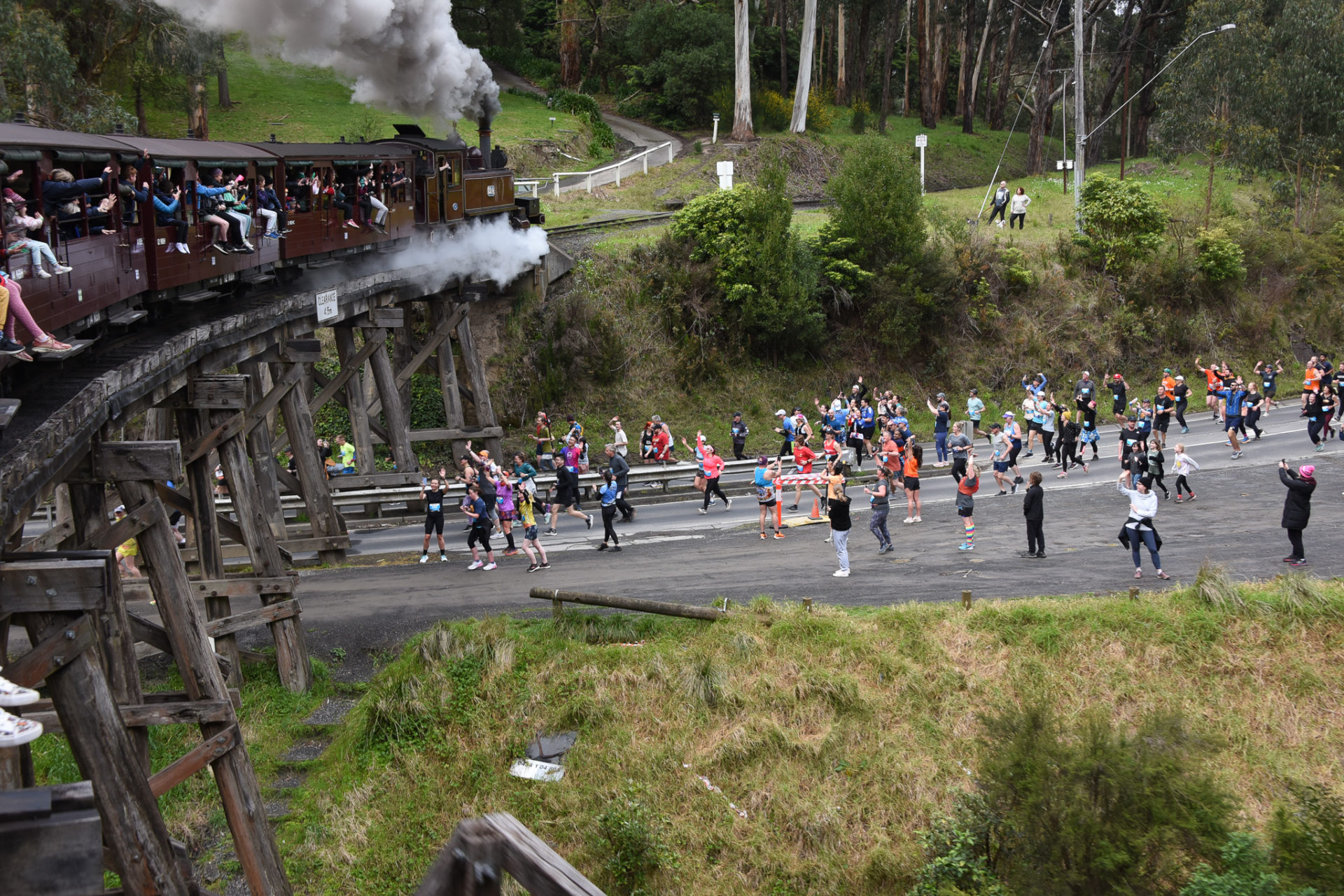 A field of runners are sprinting underneath a trestle bridge with a steam train travelling over it,