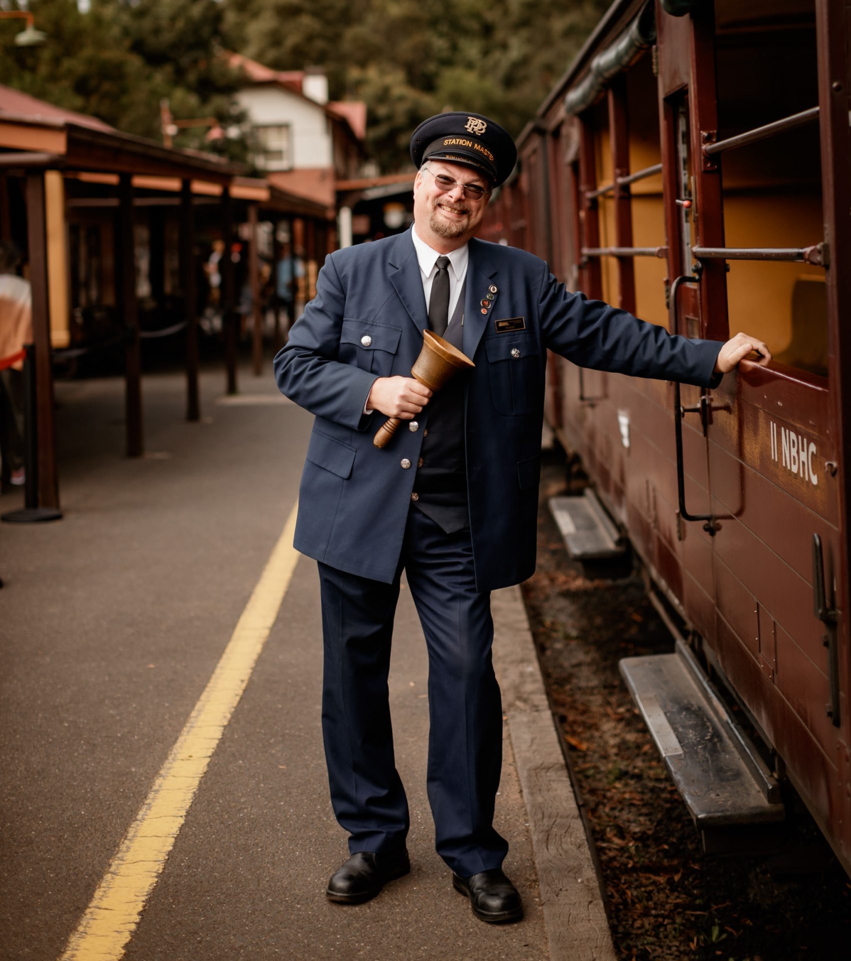 The Puffing Billy Station Master is holds onto the door handle of one of Puffing Billy's open side carriages. He is holding a bell in his right hand and is smiling.