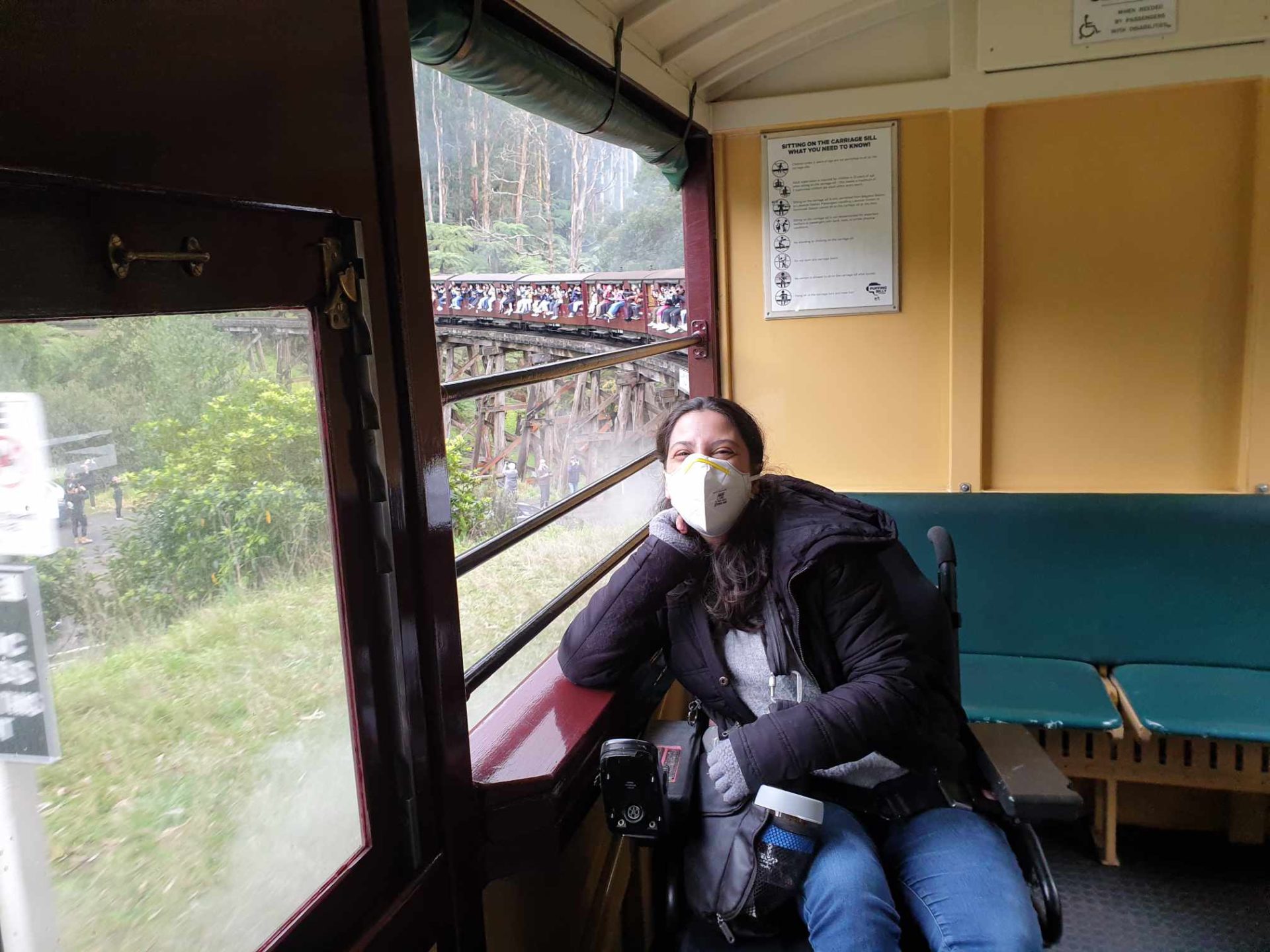 Young woman in a motorised wheelchair sits in one of Puffing Billy's accessible open side carriages. The train is travelling over a trestle bridge and green ferns can be seen outside. The young woman has warm clothes and a face mask, but you can see that she has a big smile on her face.