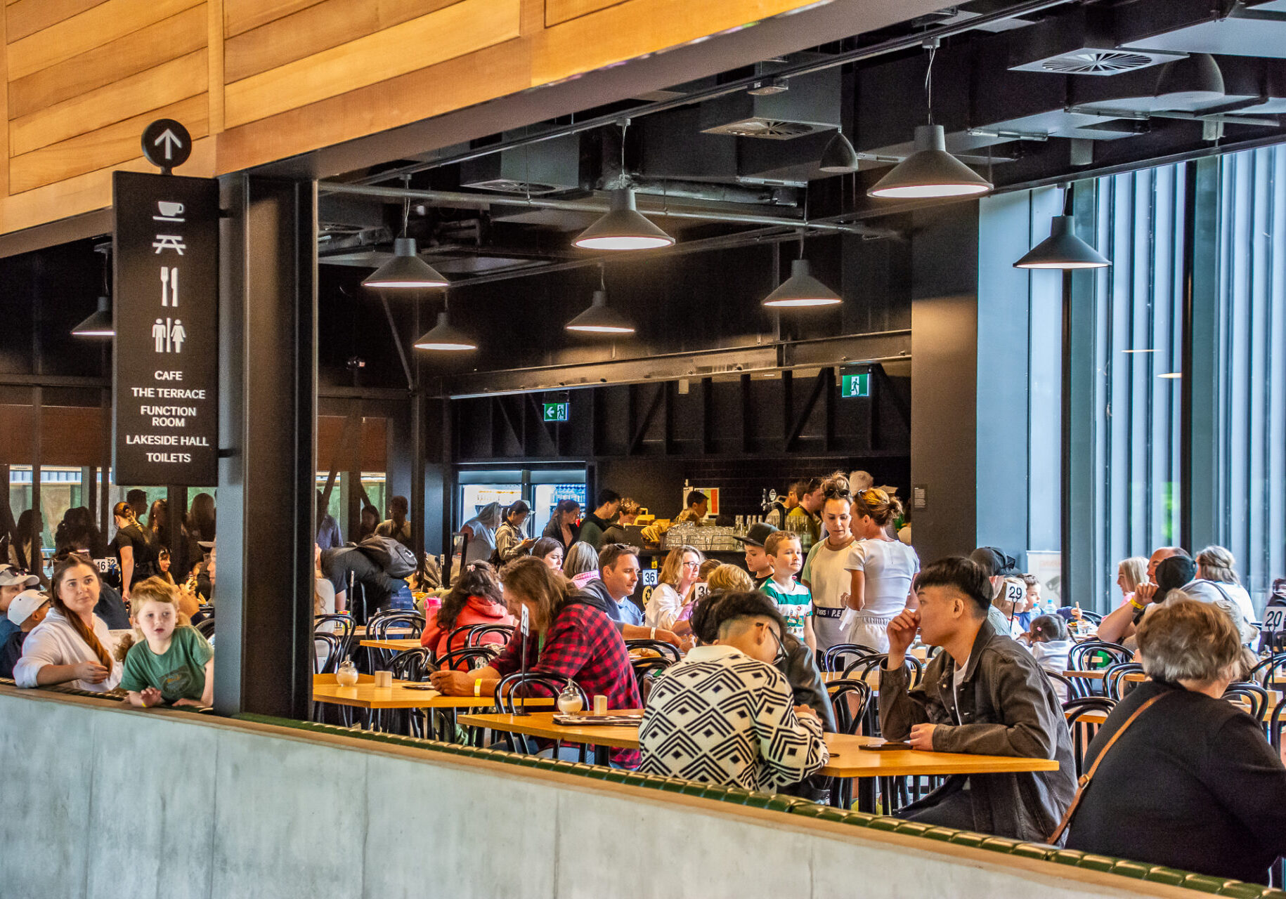 A cafe inside a building. The interior is made up of wood and black along with concrete. A large number of visitors are seen sitting at tables in the cafe.