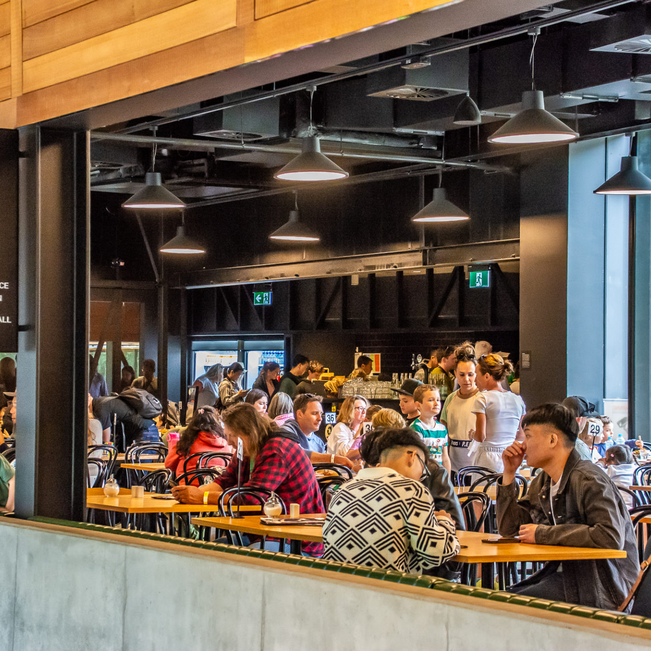 Railway Cafe with visitors sitting at tables. A cafe inside a building. The interior is made up of wood and black along with concrete. A large number of visitors are seen sitting at tables in the cafe.