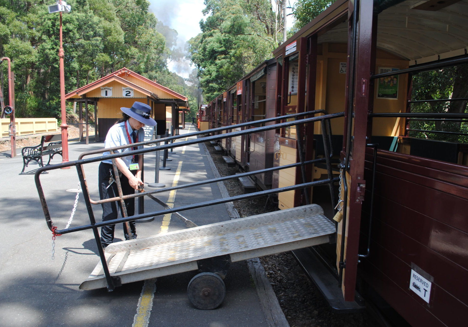 A ramp is attached to one of Puffing Billy's accessible carriages with open doors. A Puffing Billy volunteer can be seen operating the ramp to put it in place.