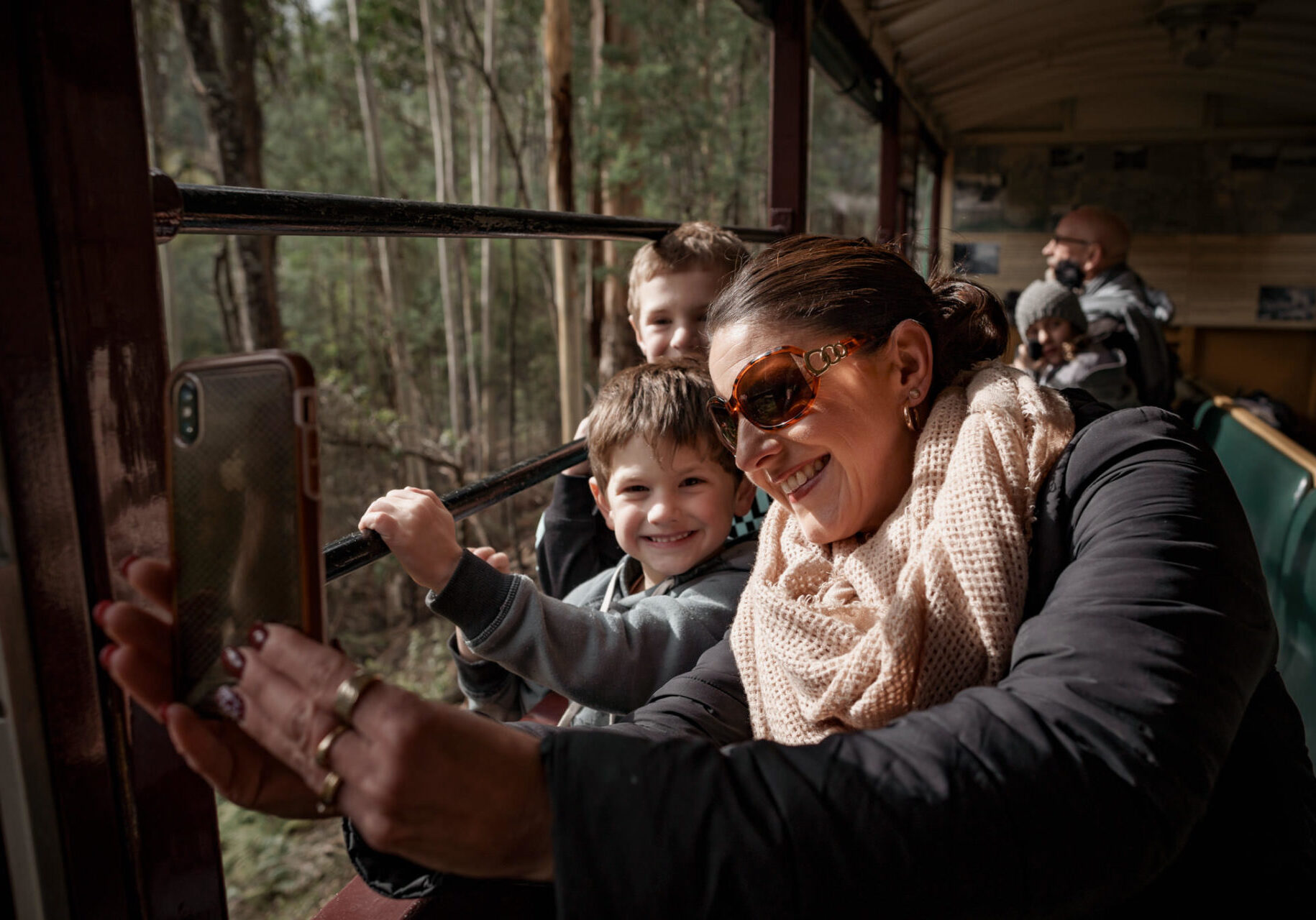 A woman and two children inside an open side train carriage. They are taking a selfie and are smiling at the camera.