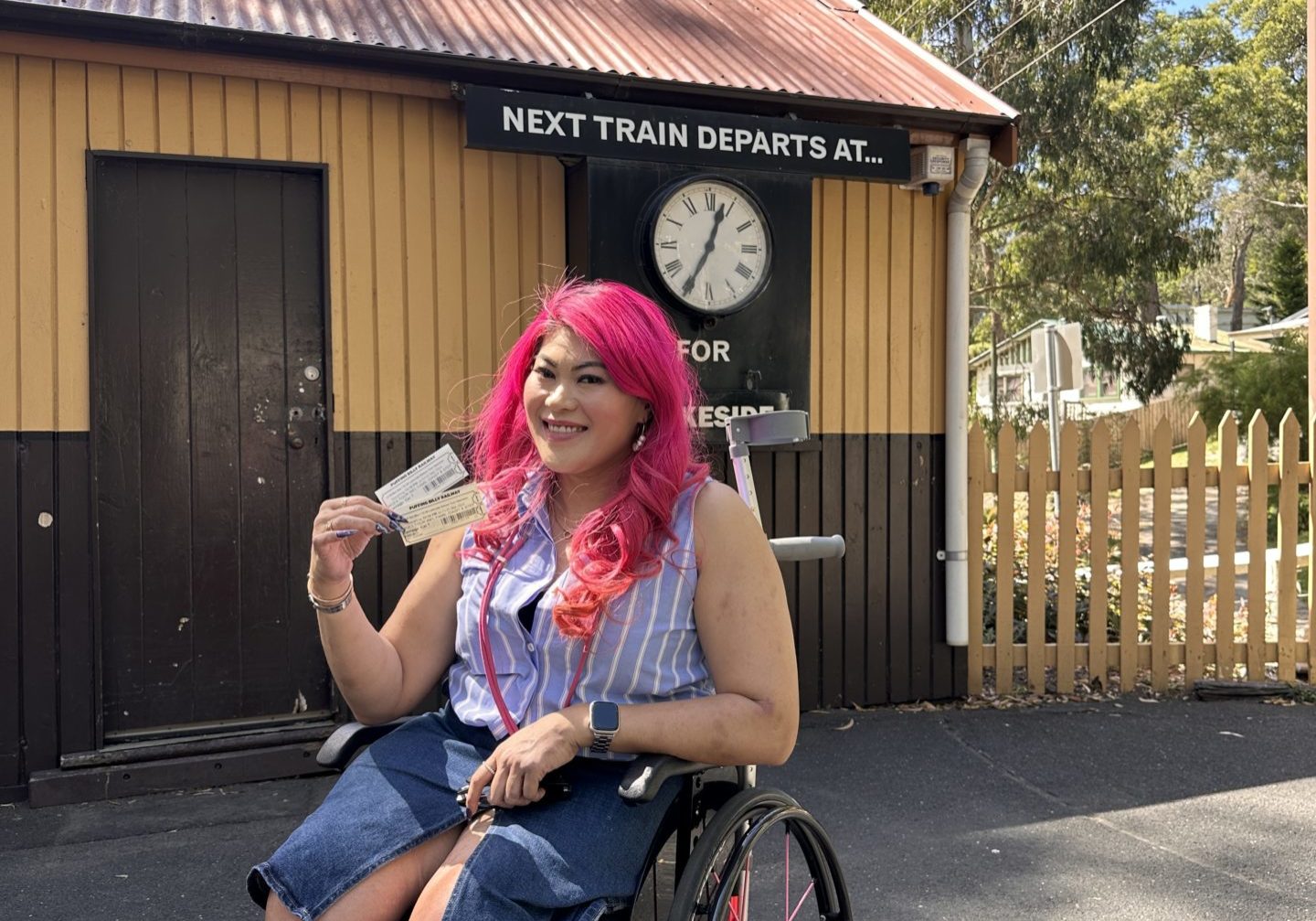 A woman in a wheelchair in front of a yellow and brown painted wooden building. An old-style clock can be seen in the background, showing the departure of the next train. The woman is holding her Puffing Billy tickets and smiling at the camera.