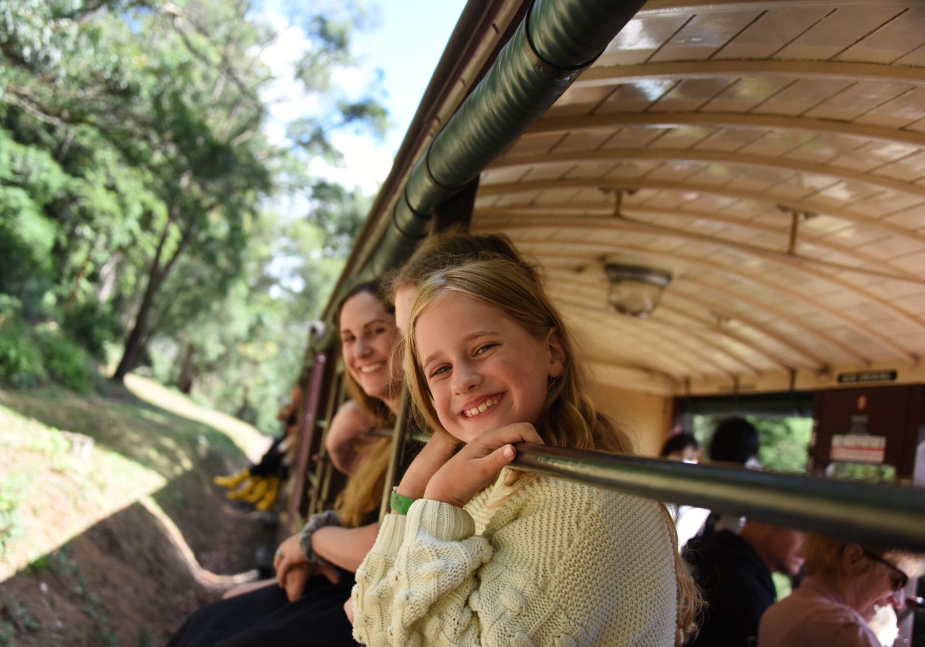 Passengers sitting with their legs dangling over the carriages sills in one of Puffing Billy's open side carriages. They are looking at the camera and smiling.