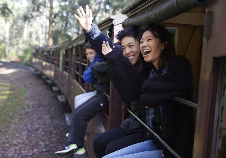 A young couple are sitting on the carriage sills of an open side carriage. They are waving and smiling to someone off-camera.