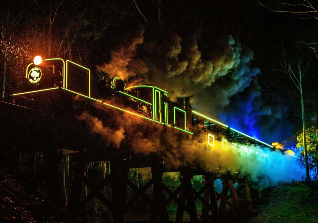 Puffing Billy Train of Lights traverses the trestle bridge over Cockatoo Creek. The locomotive and carriages are illuminated in rainbow colours.