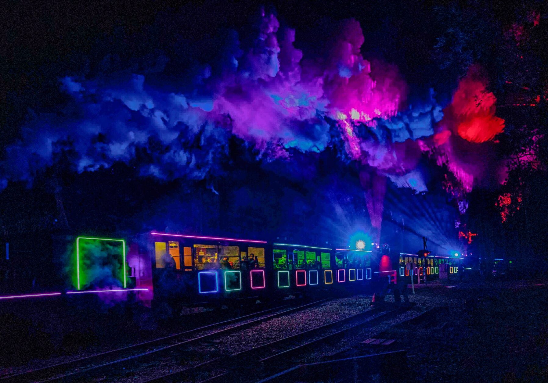 A illuminated steam train in the darkness. The train and steam are lit up in purple, pink, red, blue and green hues.