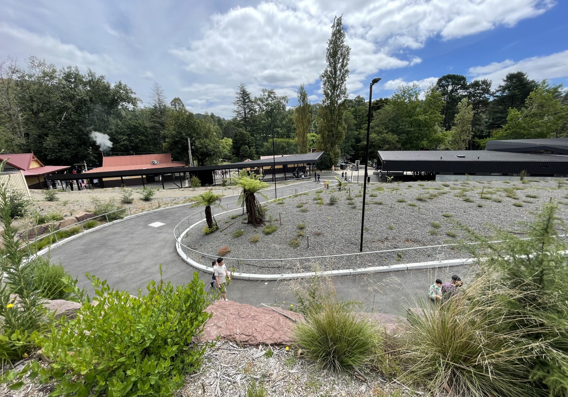 A paved pathway curves to the right down to main entrance of the Lakeside Visitor Centre. The modern centre can be seen on the right, with a black roof, while the heritage Lakeside station is on the left with warm yellow walls and red roofs. There are handrails along the path, which is on an incline.