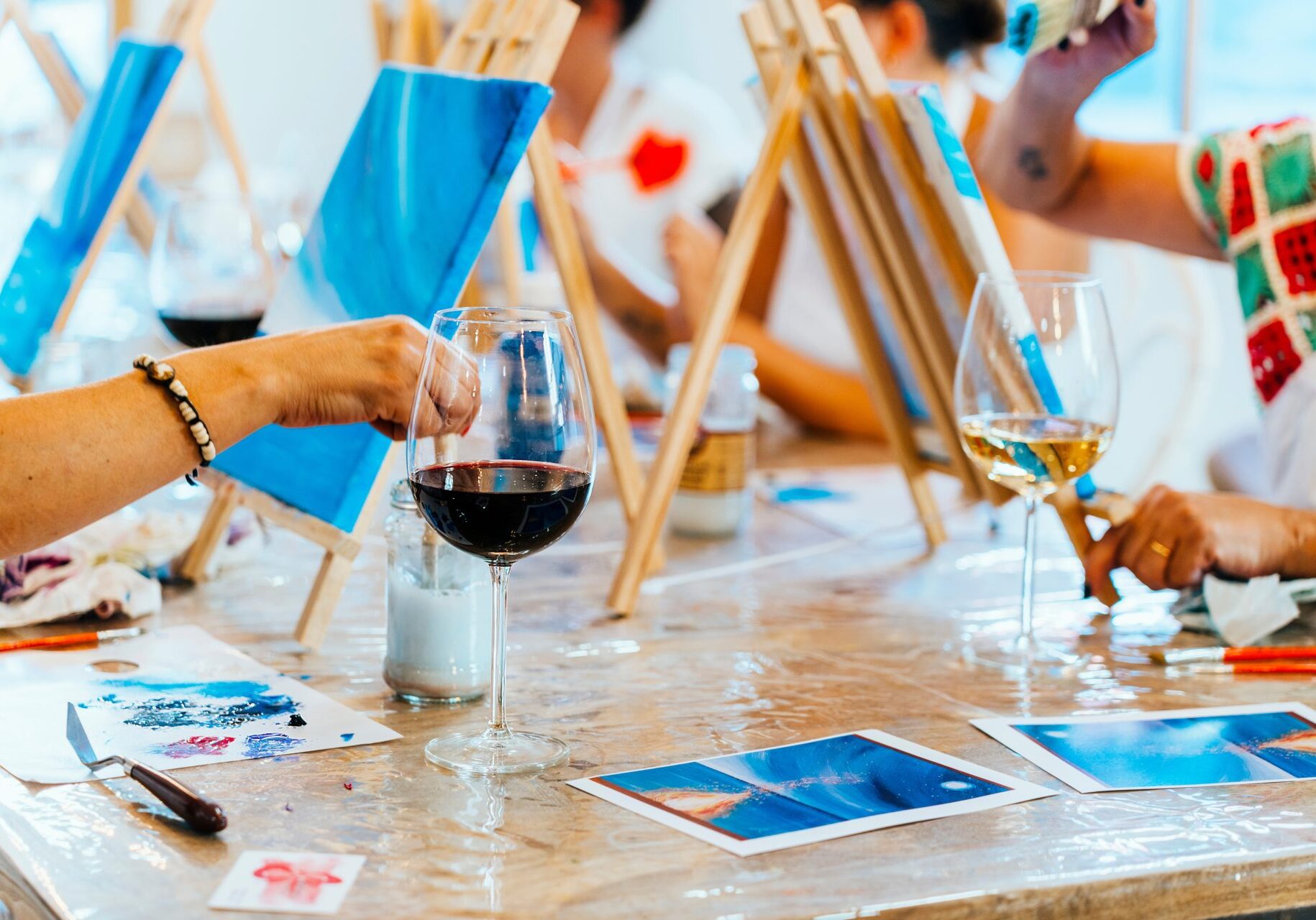 Visitors take part in a Paint and Sip Workshop. They are using wooden easels placed side by side at a table and are painting on canvases.