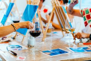 Visitors take part in a Paint and Sip Workshop. They are using wooden easels placed side by side at a table and are painting on canvases.