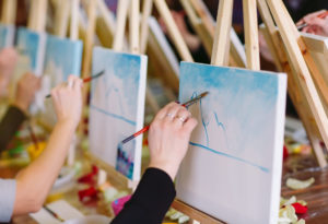 Visitors take part in a Paint and Sip Workshop. They are using wooden easels placed side by side at a table and are painting on canvases.